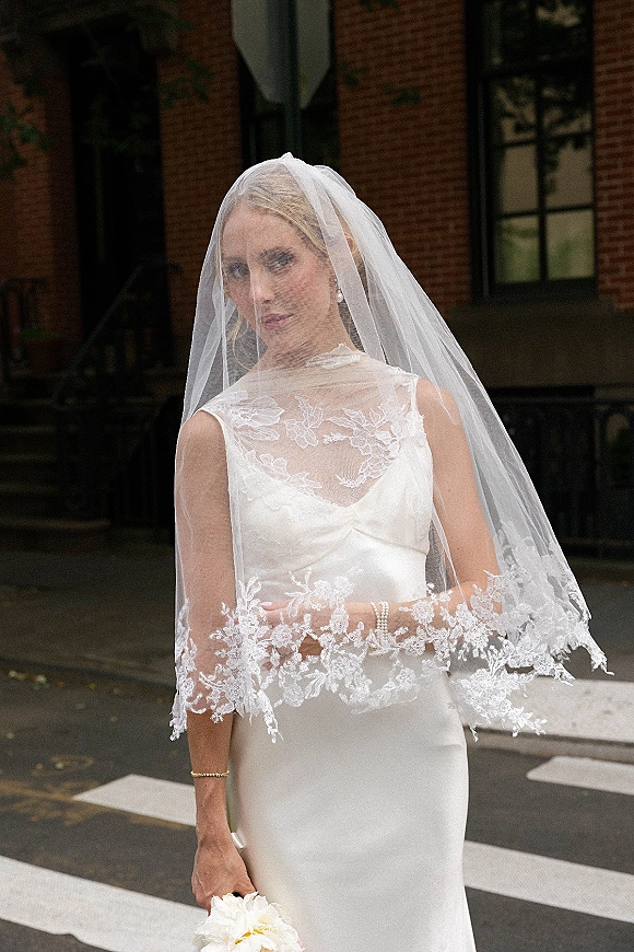 Bridal portrait of a bride in a lace wedding veil holding a bouquet, wearing pearl jewelry by a brick building with large windows on a street crosswalk