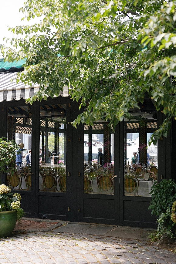 Reception venue exterior with striped awning over glass doors, bistro chairs and round tables set with white linens and floral centerpieces on a cobblestone patio