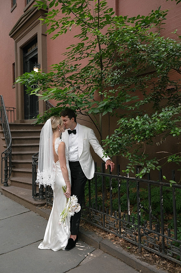 Wedding couple portrait of bride and groom kissing as she holds a bouquet on a brownstone stoop, veil trailing by wrought iron railing