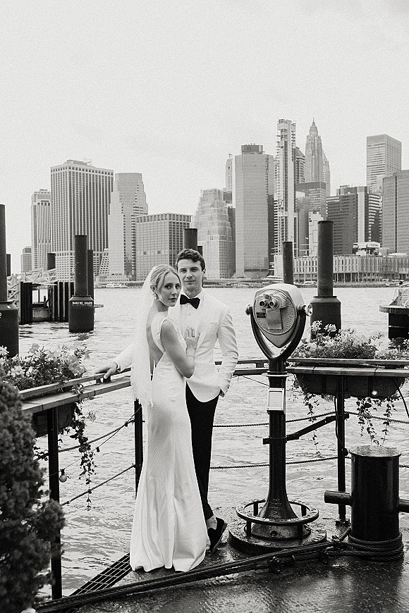 Couple portrait of bride in veil and backless wedding dress with groom in white tuxedo and black bow tie by waterfront skyline pier railing