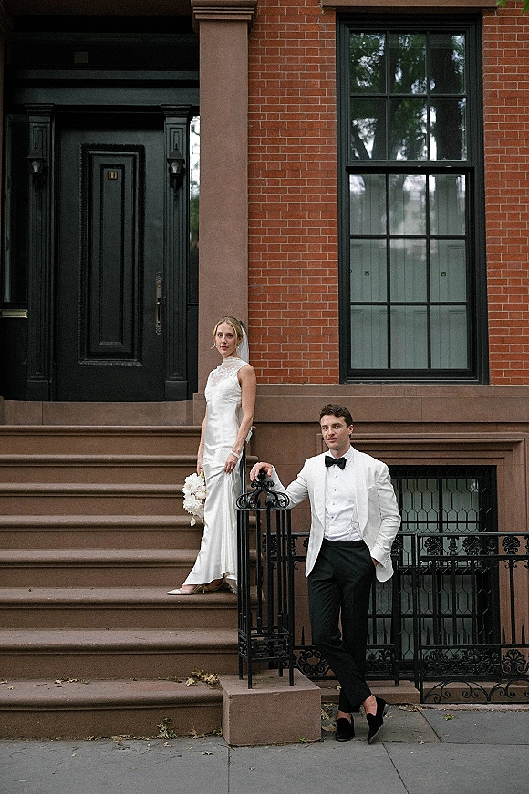 Couple portrait of bride in sleek satin dress and veil holding a white bouquet beside groom in white tuxedo by a brick brownstone door