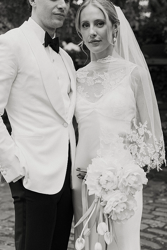 Couple portrait in a black and white wedding portrait, bride in lace dress and veil holding a ribboned bouquet beside groom in white jacket on a garden path