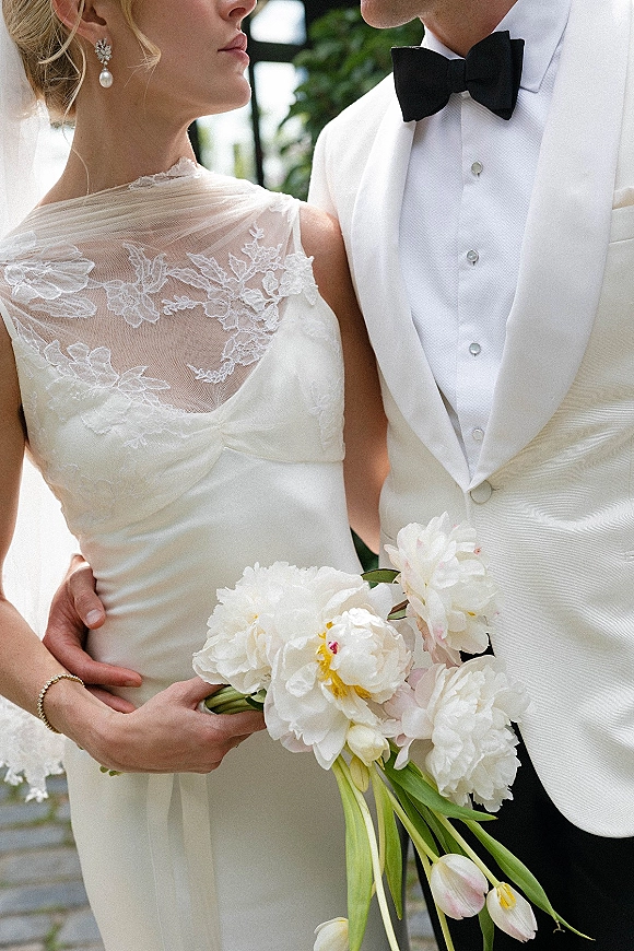 Couple portrait of bride and groom close up, embracing on stone steps with greenery, her white peony bouquet and lace neckline visible