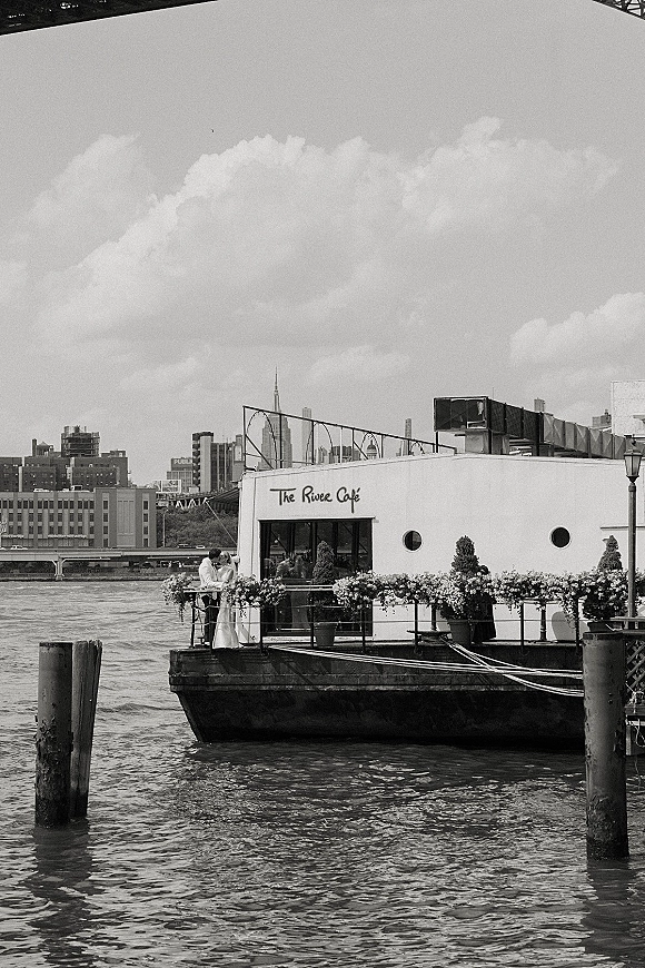 Couple portrait in a black and white wedding portrait style, bride in long sleeve dress and groom in suit by a railing with river and city skyline behind