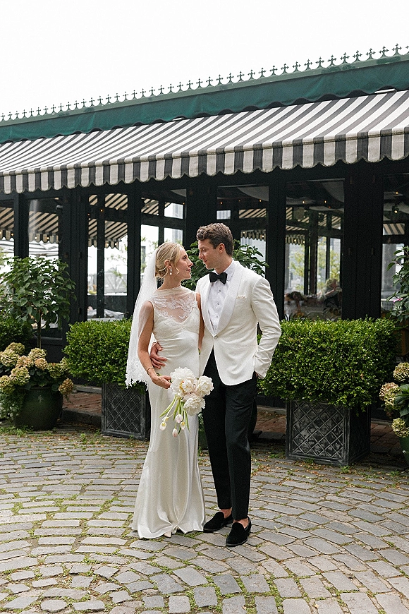 Couple portrait of bride in lace high-neck dress and veil with white bouquet, groom in white jacket and bow tie in cobblestone courtyard