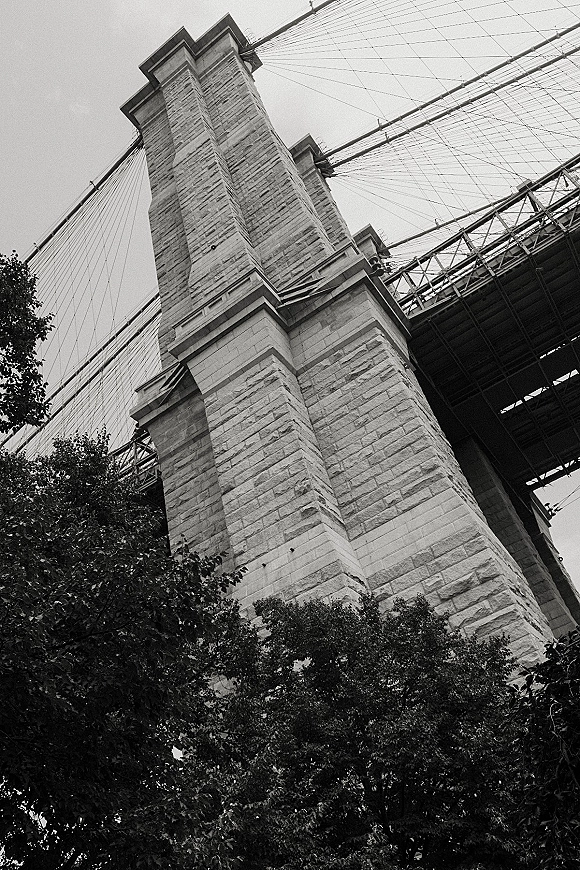 Bridge architecture in black and white, showing a stone tower with suspension cables and steel beams from a low angle, trees and sky behind