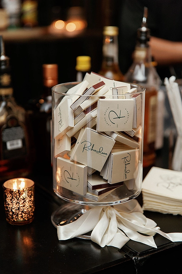 Wedding favors with custom matchboxes wedding favors in a glass cylinder jar tied with a satin ribbon bow beside a votive candle on a dim bar counter