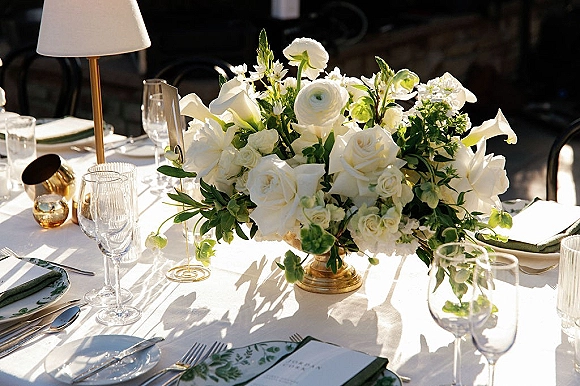 Reception tablescape with wedding table centerpiece of white flowers and greenery in a gold compote, set with menus, glasses, and lamp on a sunlit patio