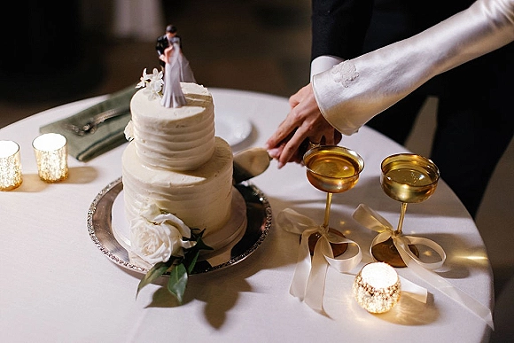 Wedding cake cutting with bride and groom cutting cake, hands on knife beside champagne coupes on a dimly lit reception table with white roses