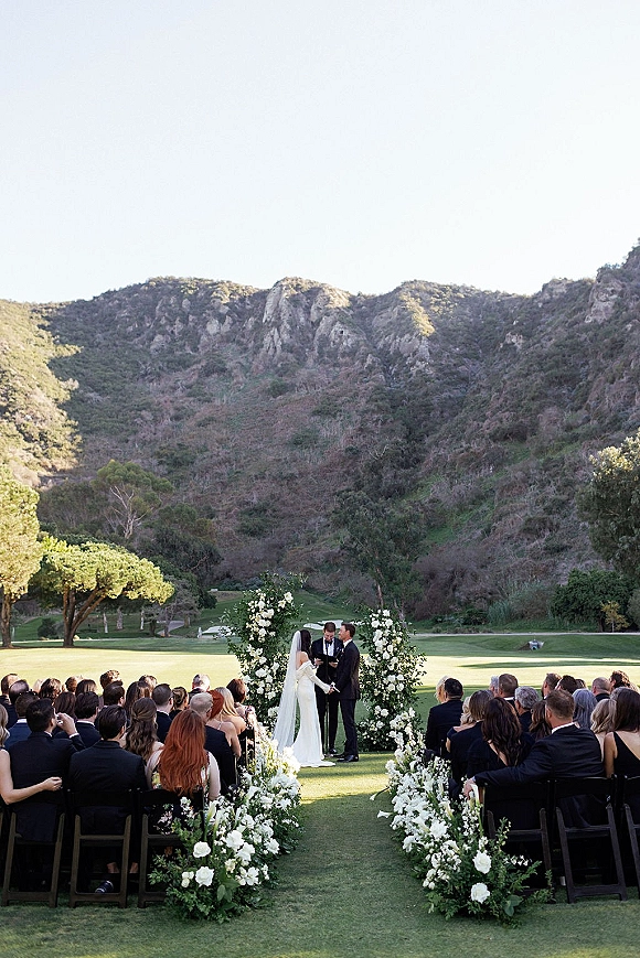 Wedding ceremony with bride and groom under a white floral arch, veil flowing, guests seated on a lawn with mountain backdrop