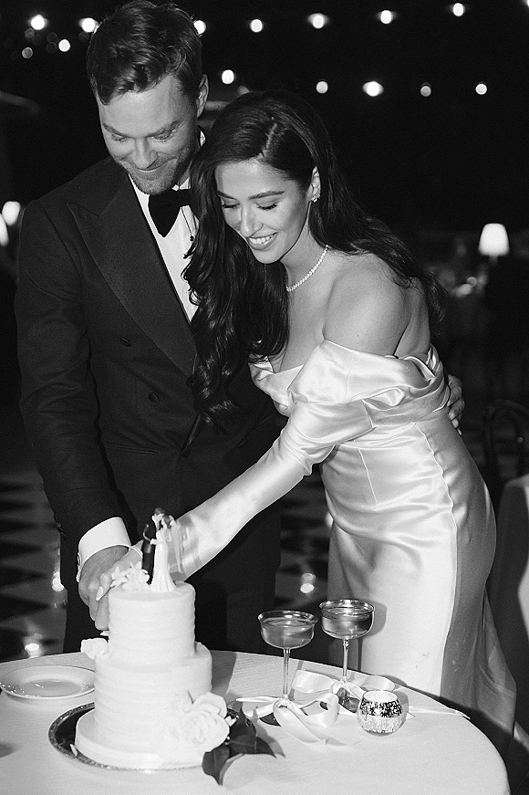 Cake cutting as bride and groom slice a white buttercream wedding cake in a dark reception with string lights and champagne coupes