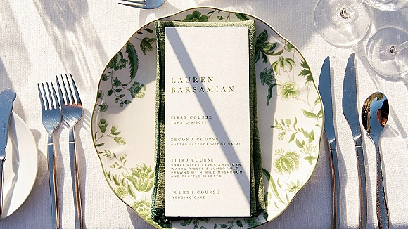 Wedding menu card resting on a patterned dinner plate with green napkin, silver flatware, and water and wine glasses on a white tablecloth