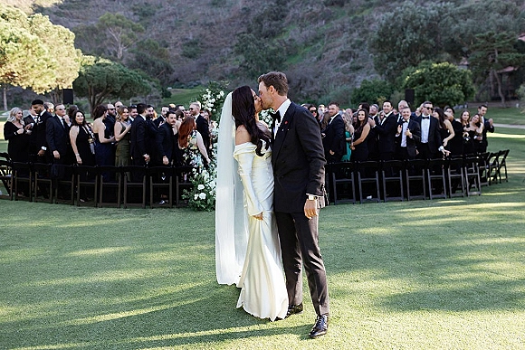 Wedding kiss as bride in off shoulder dress and veil kisses groom in tuxedo, with guests and ceremony chairs on a hillside lawn