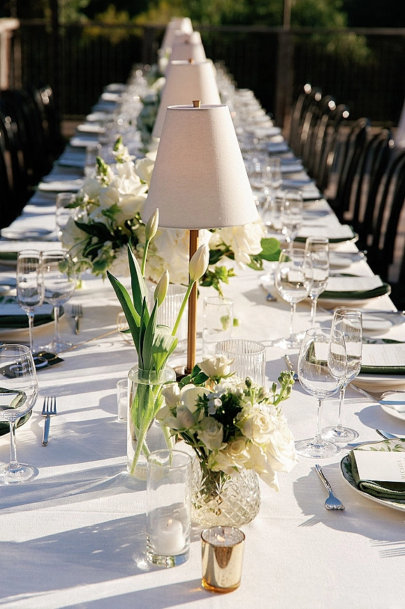 Reception tablescape with a long banquet table set in white linens, tulip and rose florals, greenery garland, and candlelight on a sunny patio