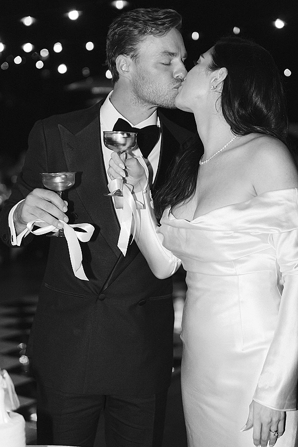 Wedding kiss portrait of newlyweds raising coupe glasses, bride in satin dress and jewelry, groom in tuxedo under string lights at night