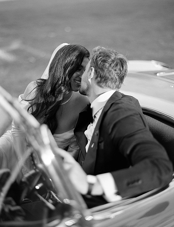 Wedding couple portrait in a vintage car, bride in veil and pearl necklace leaning close to groom in tuxedo, blurred street behind