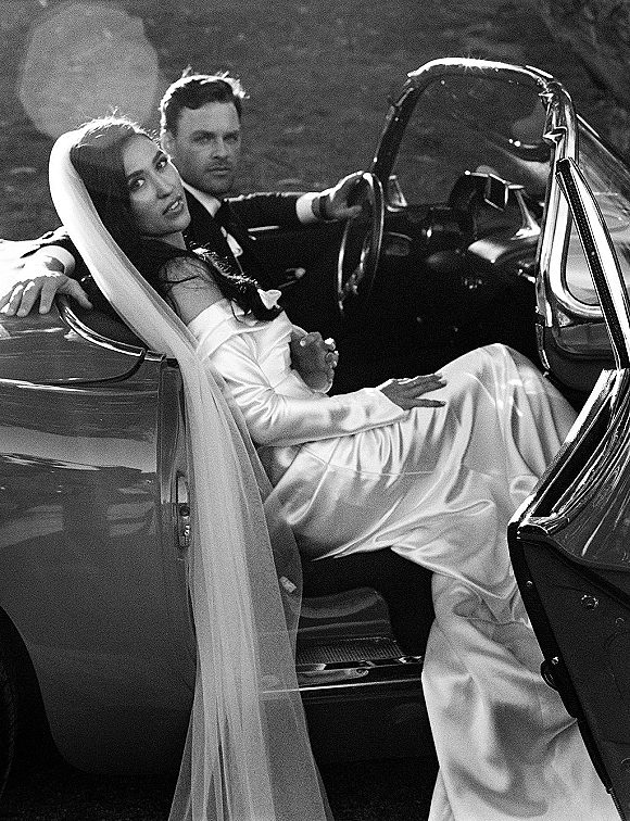 Couple portrait of bride and groom in car, in a classic convertible, her long veil flowing as he drives down a foliage-lined road