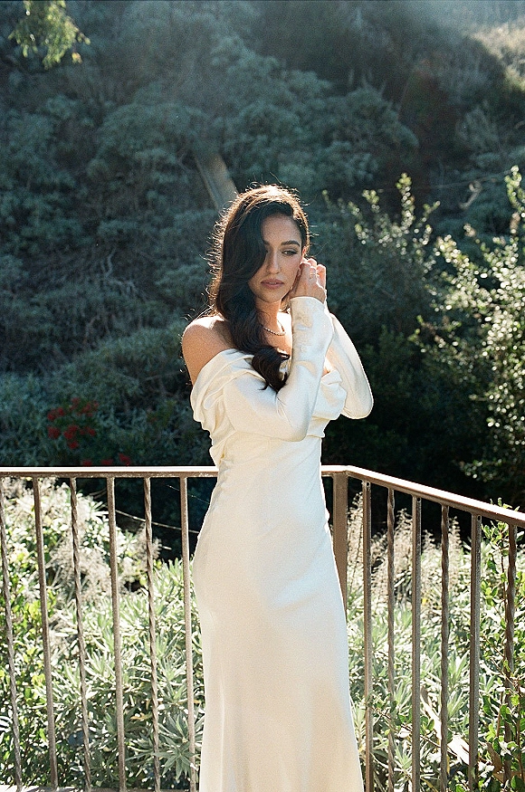 Bridal portrait of a bride in an off the shoulder wedding dress, touching her earring on a sunlit balcony with green hillside behind