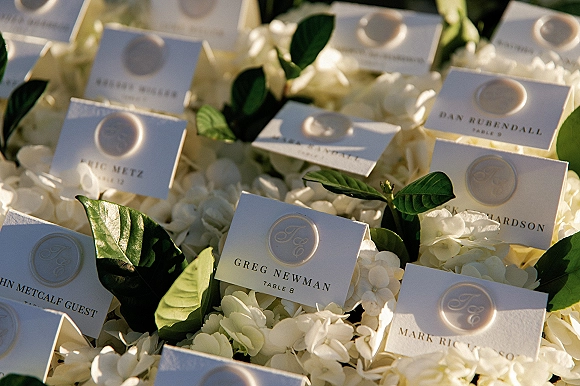 Escort cards in a wedding escort card display, finished with monogram wax seals, nestled on white florals and greenery in soft sunlight