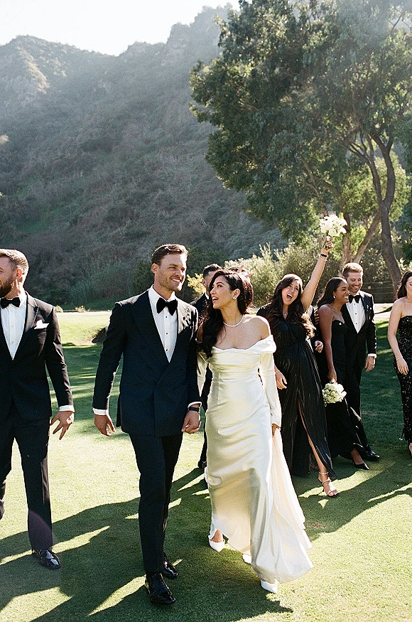 Wedding party portrait with bride in a wedding dress and groom in tuxedo, bridesmaids in black, on a sunlit lawn with mountains behind