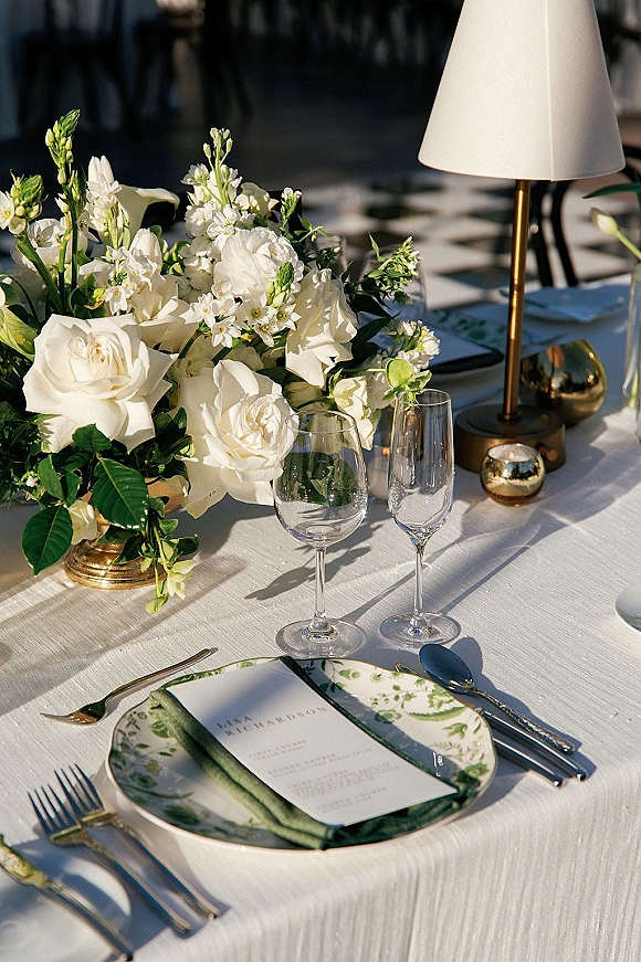 Reception tablescape with a wedding place setting, white rose centerpiece in a gold vase, green napkin, menu card, and candlelit lamp outdoors