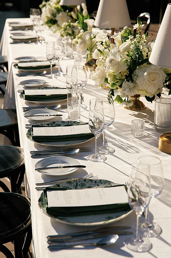 Reception tablescape wedding table setting with white rose and tulip centerpieces, taper candles, lamps, and green napkins on a sunlit patio