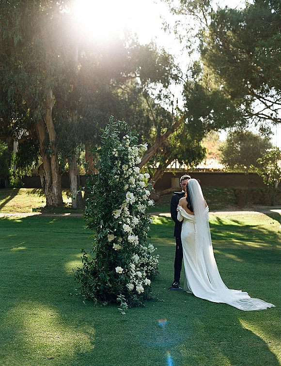 Ceremony moment as bride and groom at altar exchange vows, her long veil trailing beside white flowers and greenery in sunlit garden lawn