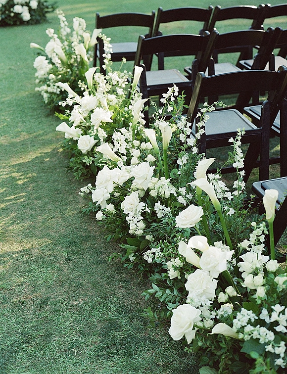 Ceremony aisle flowers in a wedding aisle flower arrangement of white calla lilies and roses with greenery garland beside black chairs on grass lawn