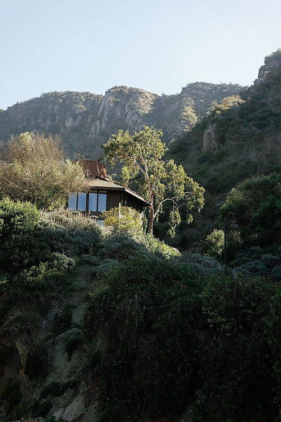 Mountain cabin exterior with large windows and metal roof, balcony railing and wall sconces, set on a wooded hillside under blue sky