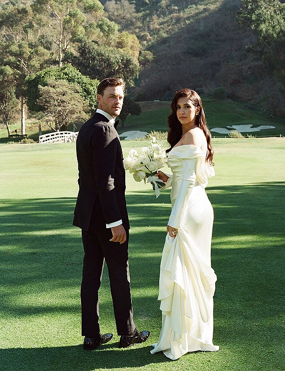Couple portrait of bride and groom outdoor portrait, she holds a white bouquet in an off-shoulder dress on a golf course lawn with bridge