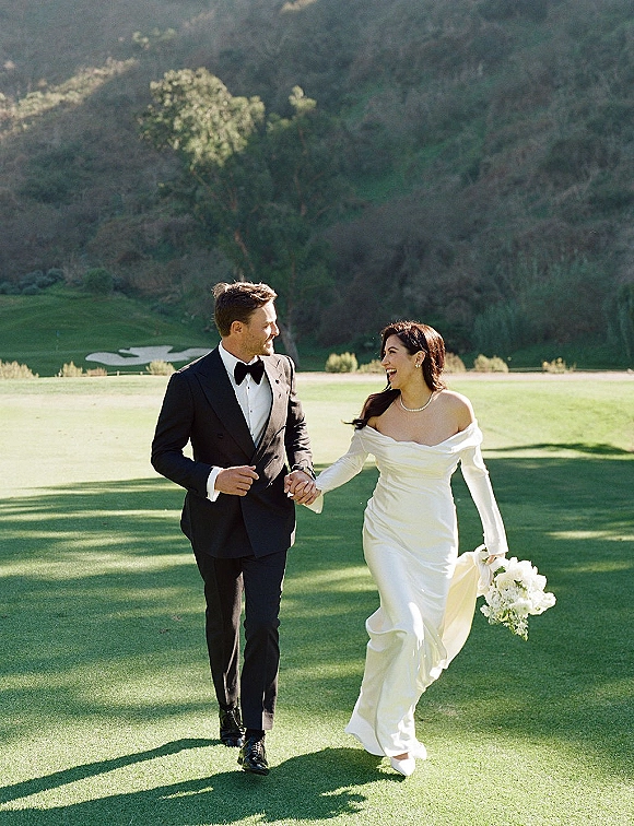 Couple portrait of bride and groom walking hand in hand on a golf course lawn, bride holding a bouquet in a strapless gown, groom in black tuxedo