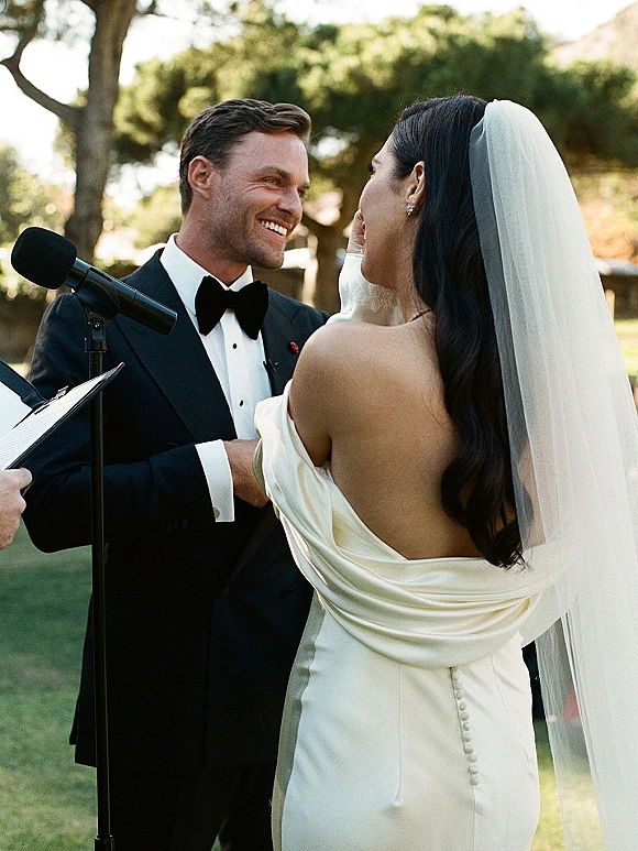 Wedding vows as bride in veil and off-the-shoulder gown reads from papers at microphone, groom in black bow tie smiles outdoors