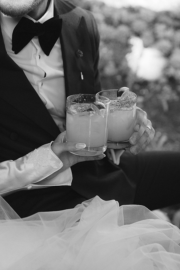 Wedding drinks toast as bride and groom in tuxedo and lace sleeve clink salted-rim cocktails with lime wedges against greenery