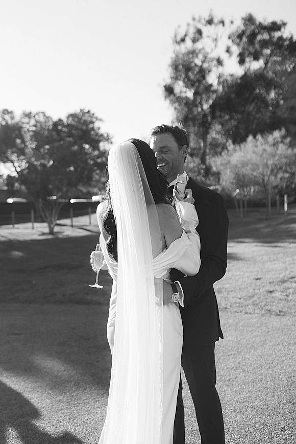 Couple portrait in a black and white wedding portrait style, bride in veil and dress embracing groom in suit on a sunlit lawn with trees