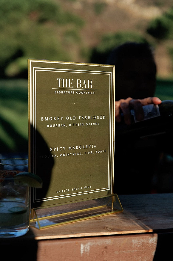 Bar menu sign wedding bar sign in an acrylic holder on a wooden bar top beside a cocktail glass with lime, greenery behind