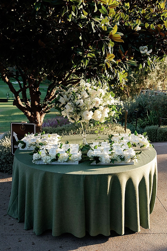 Escort card table with wedding escort cards on a sage green linen tablecloth, white florals and greenery garland in a garden setting