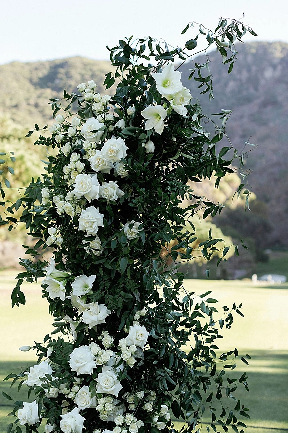 Wedding ceremony arch with an asymmetrical floral arch of white roses and lilies, lush greenery, set on a hillside lawn with mountains beyond