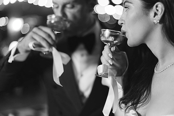 Wedding toast as bride and groom clink champagne coupe glasses with ribbon accents, tuxedo and bridal jewelry glowing under bokeh string lights