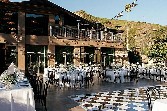Outdoor reception setup with round tables in white linens, black bistro chairs, floral centerpieces, and checkered dance floor under string lights