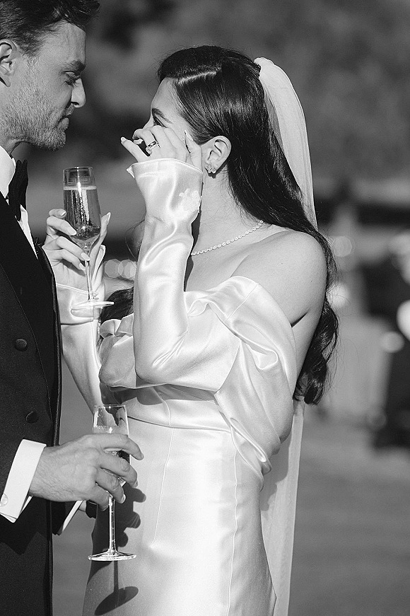 Wedding couple portrait in black and white, bride wiping tears as they toast with champagne flutes in sunlight with a blurred crowd behind