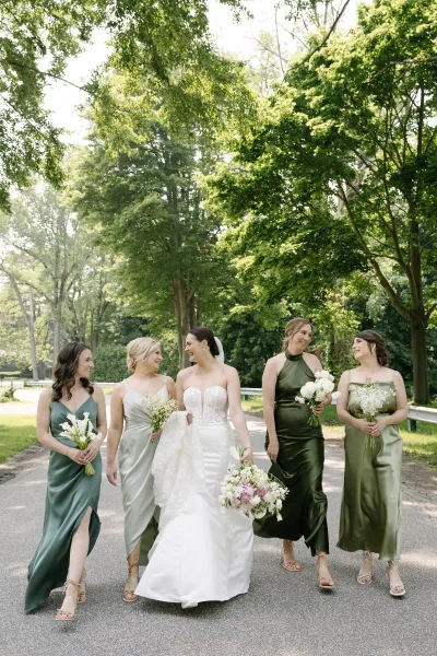 Bride with bridesmaids walking in sage green dresses, bride in lace gown and veil holding bouquets on a sunlit tree-lined road