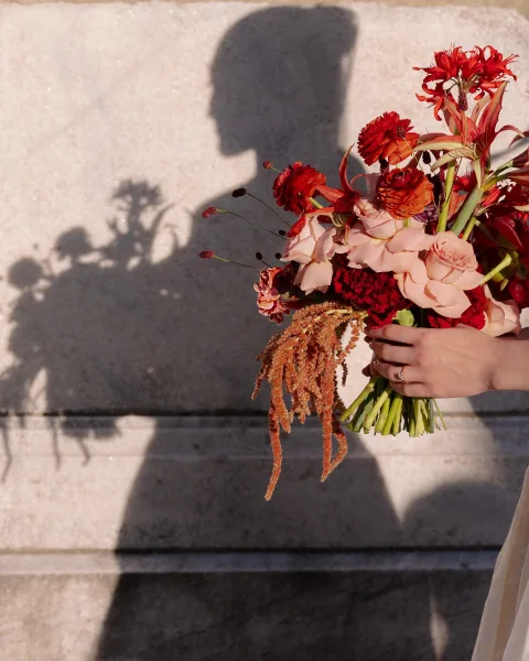 Bridal bouquet with red wedding bouquet blooms in a bride’s hand, engagement ring shining, against a stone wall and shadow silhouette