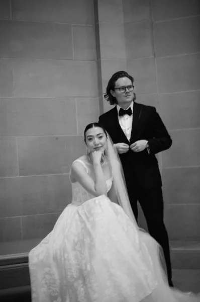 Couple portrait in a black and white wedding portrait style, bride seated in veil and gown as groom in tuxedo stands by a stone wall indoors