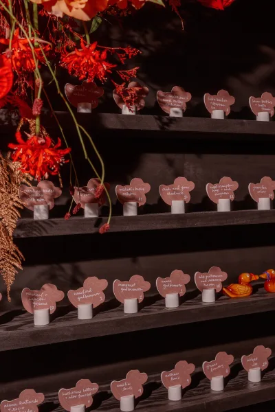 Escort card display with wedding escort cards on black shelving, heart-shaped calligraphy place cards amid red florals, greenery, and ducks
