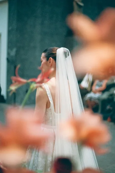 Bridal portrait of a bride in veil and sleeveless gown, seen from behind with long veil trailing in a garden aisle near a stone wall