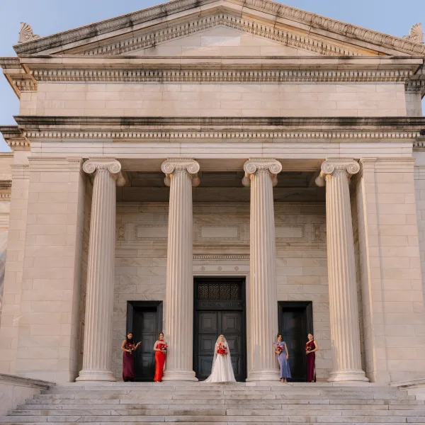 Bridal party portrait with bride and bridesmaids on stone steps, bouquets in hand, framed by marble columns and blue sky