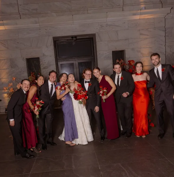 Wedding party portrait with bride and groom with wedding party laughing in an uplit entryway, holding bouquets with bright red florals