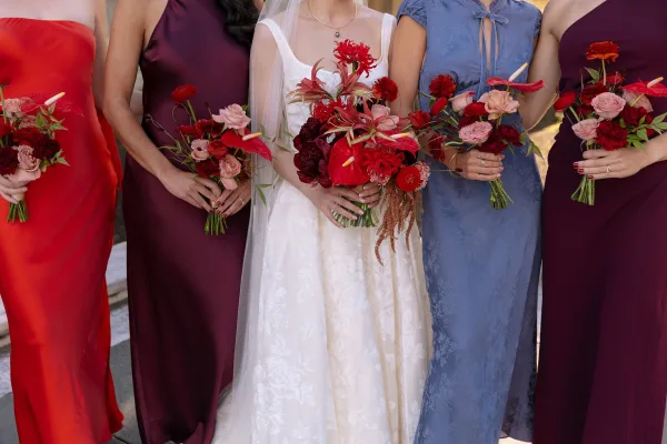Bridesmaid bouquets in red and burgundy bouquets with anthurium and roses, held beside a bridal gown and veil on stone steps outdoors