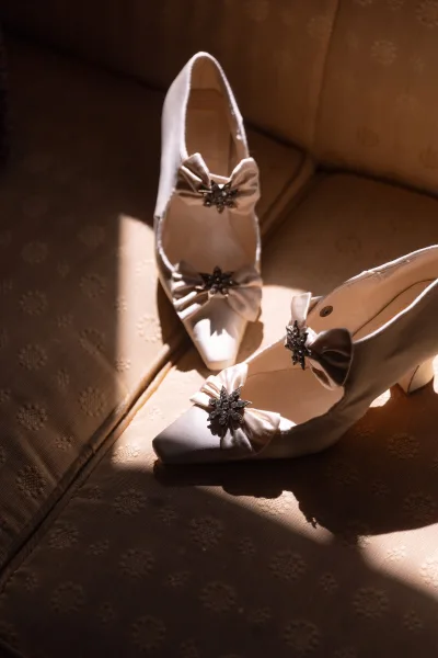 Bridal shoes with satin bows and crystal starburst brooches, wedding heels detail resting on an upholstered chair in warm sunlight