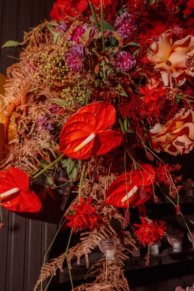 Wedding floral arrangement with red anthurium bouquet, peach roses and purple blooms in a glass vase on a tabletop against a dark wood wall